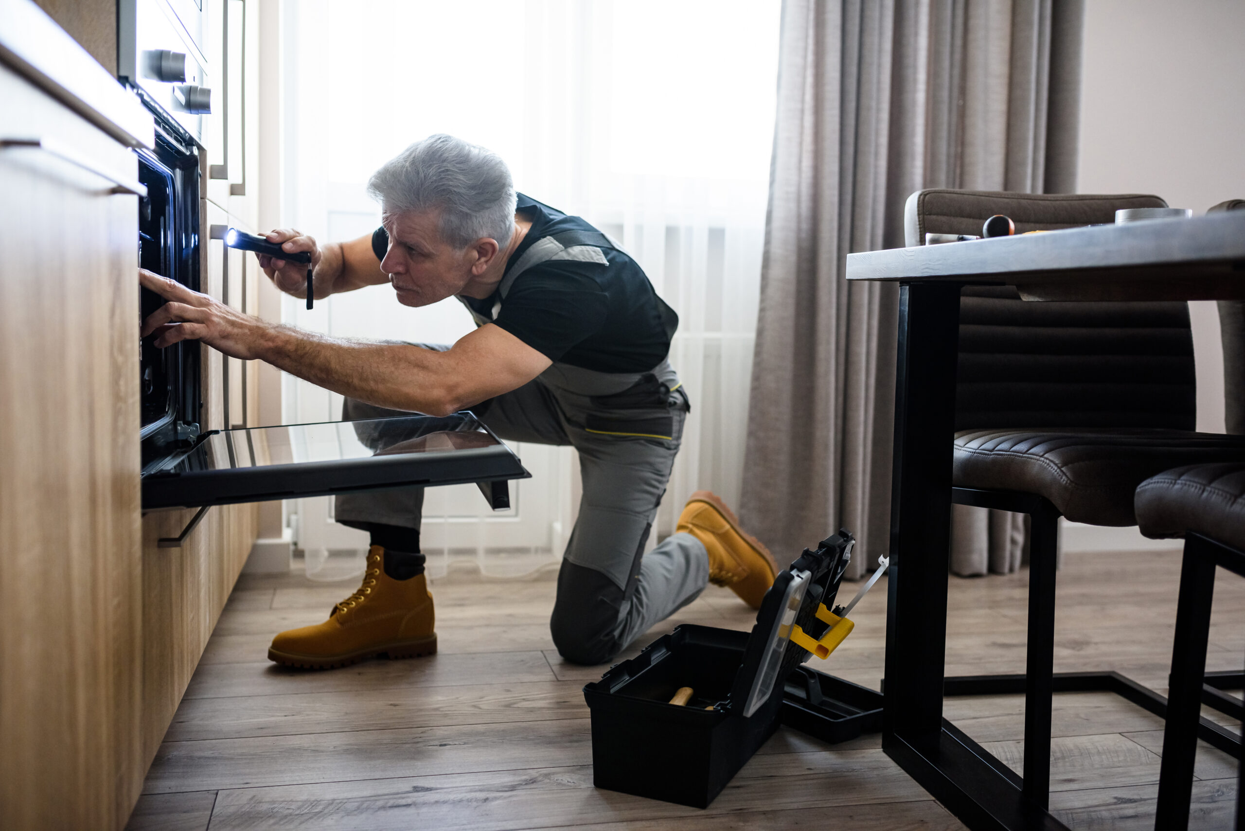 Full length shot of aged repairman in uniform working, examining broken oven in the kitchen using flashlight. Repair service concept. Focus on person. Side view. Horizontal shot